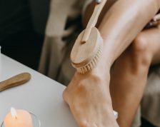 Dry brushing body brush for exfoliating dry skin, lymphatic drainage and cellulite treatment. Woman brushing skin leg with dry wooden brush to prevent cellulite and body problem in bedroom at home