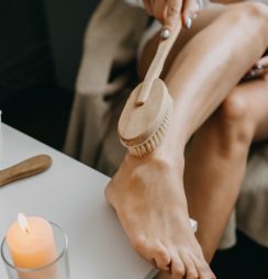 Dry brushing body brush for exfoliating dry skin, lymphatic drainage and cellulite treatment. Woman brushing skin leg with dry wooden brush to prevent cellulite and body problem in bedroom at home