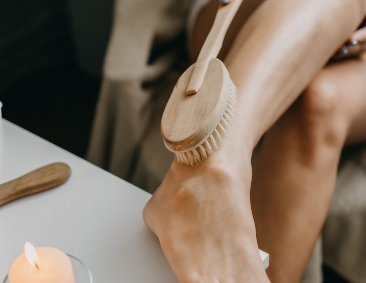 Dry brushing body brush for exfoliating dry skin, lymphatic drainage and cellulite treatment. Woman brushing skin leg with dry wooden brush to prevent cellulite and body problem in bedroom at home